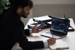 Smart focused Indian broker investor, stock trader using smartphone and laptop to analyze and invert in the financial market of cryptocurrencies, buy or sell crypto coins, planning strategy