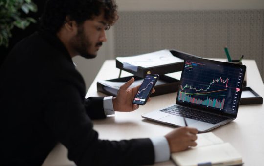 Smart focused Indian broker investor, stock trader using smartphone and laptop to analyze and invert in the financial market of cryptocurrencies, buy or sell crypto coins, planning strategy